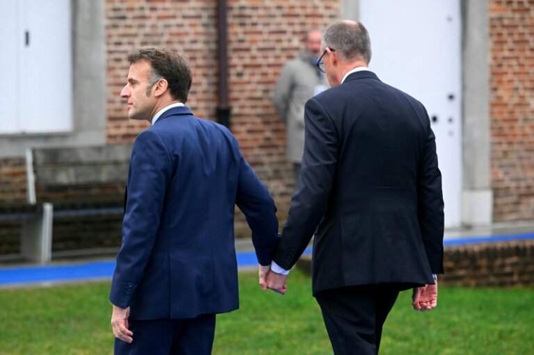 Emmanuel Macron (G) et Friedrich Merz se tiennent la main à leur arrivée au sommet informel de l'UE au château belge d'Alden Biesen en Belgique le 12 février 2026 © NICOLAS TUCAT