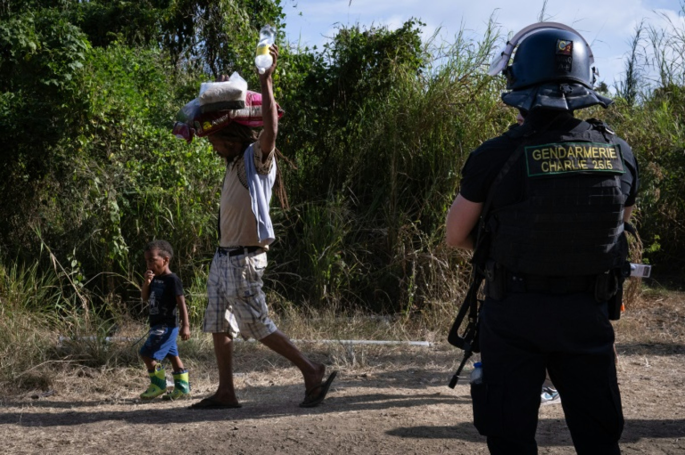 Un habitant, transportant sur sa tête des sacs de provisions, passe avec des enfants, devant un gendarme à un barrage routier à Saint-Louis, au sud de Nouméa, le 23 septembre 2024 en Nouvelle-Calédonie © SEBASTIEN BOZON