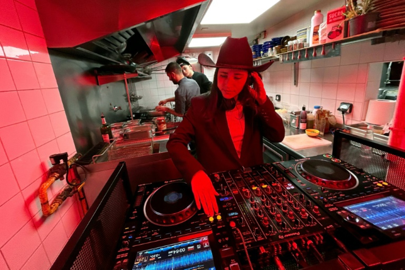 La DJ Carla Mo (c) dans les cuisine d'un restaurant lors du spectacle musical "Let Him Cook" diffusé sur les médias sociaux, pendant que le cuisinier Enzo Franceschi (g) prépare un plat sous le regard du fondateur du spectacle Théo Ferrato (d), le 9 décembre 2024 à Marseille © Viken KANTARCI