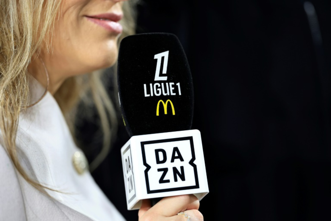 Une journaliste avec un icro portant les logos de DAZN et de la Ligue 1 le 28 février 2025 au stade Louis-II avant un match de championnat entre Monaco et Reims. La médiation dans le litige financier sur les droits TV entre le diffuseur principal de la L1, DAZN, et la Ligue de football professionnel (LFP) prend fin mardi © Valery HACHE