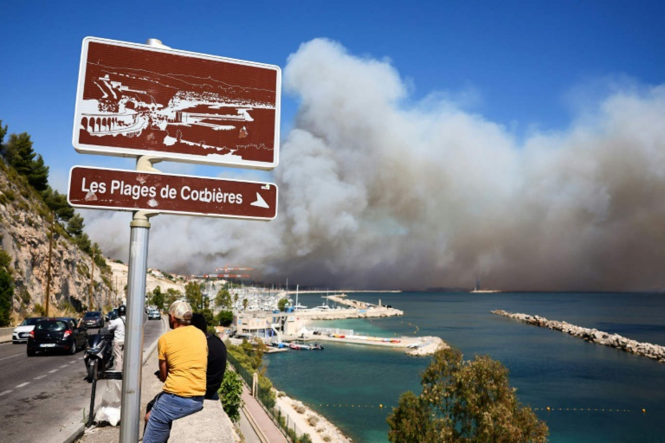 la plage des Corbières, à Marseille, dans le sud de la France, le 8 juillet 2025, des passants observent la fumée d'un incendie de forêt qui fait rage en arrière-plan © Clement MAHOUDEAU