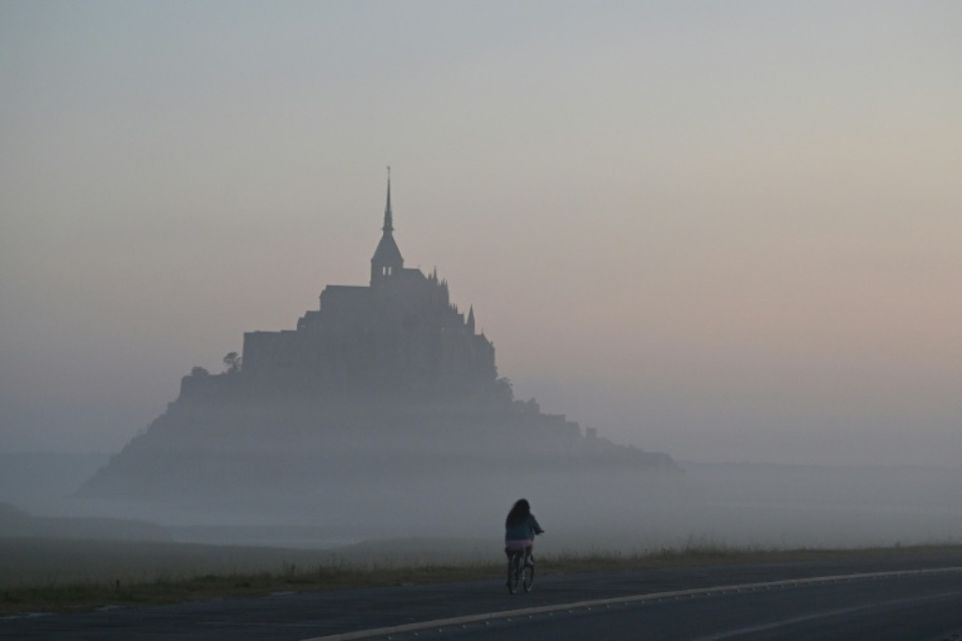 Un cycliste passe devant le Mont-Saint-Michel, enveloppé par la brume matinale, le 9 juillet 2025 © Damien MEYER